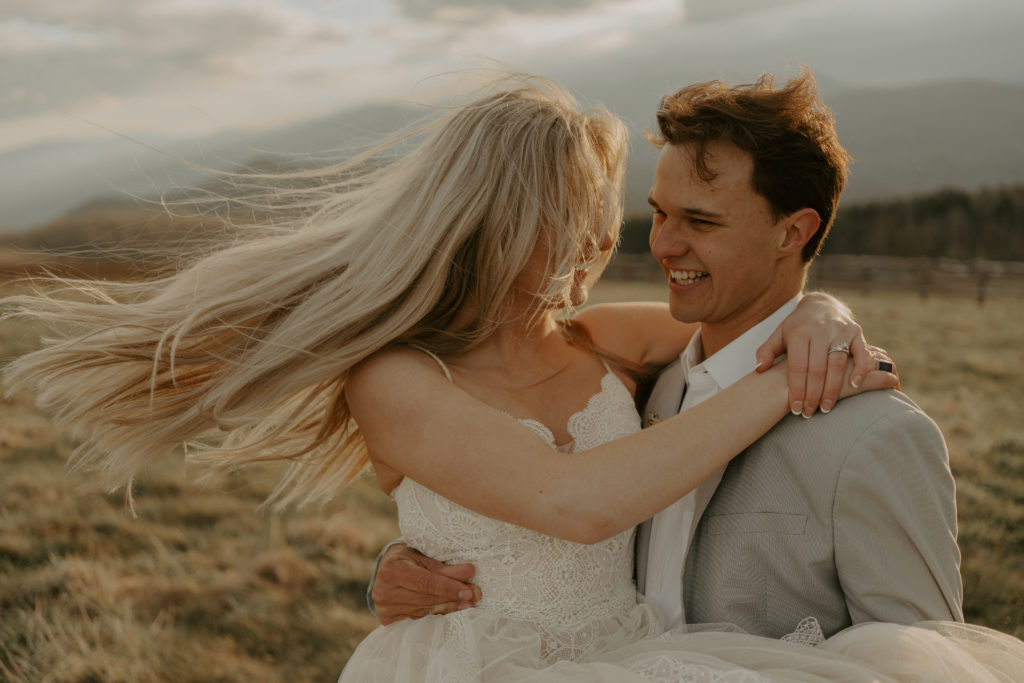 A man is holding a woman in his arms while her hair is flowing in the wind and they are dressed in their wedding attire during their Asheville mountain elopement.