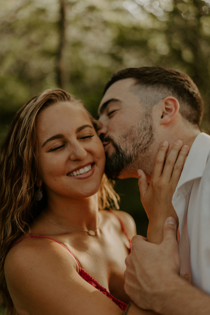 A man kisses a womans cheek as she smiles during their Asheville mountain engagement.