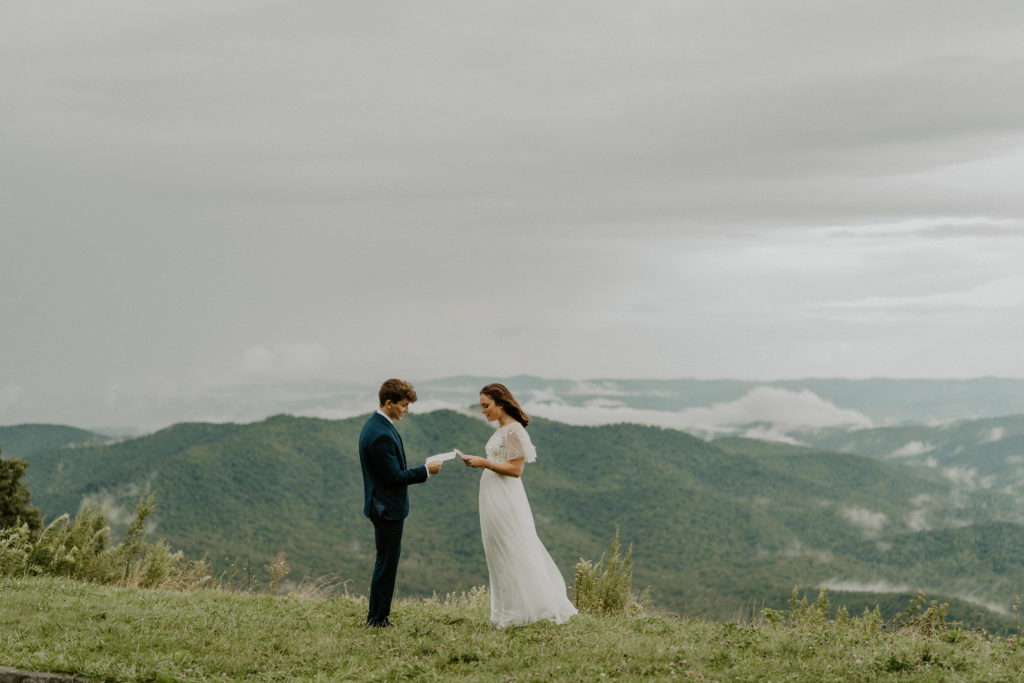 man and woman facing each other while reading wedding vows. The Blue Ridge mountains are behind them, it is overcast and small low clouds can be seen around the mountain tops. The woman is wearing a wedding dress and the man is wearing a navy suit. 