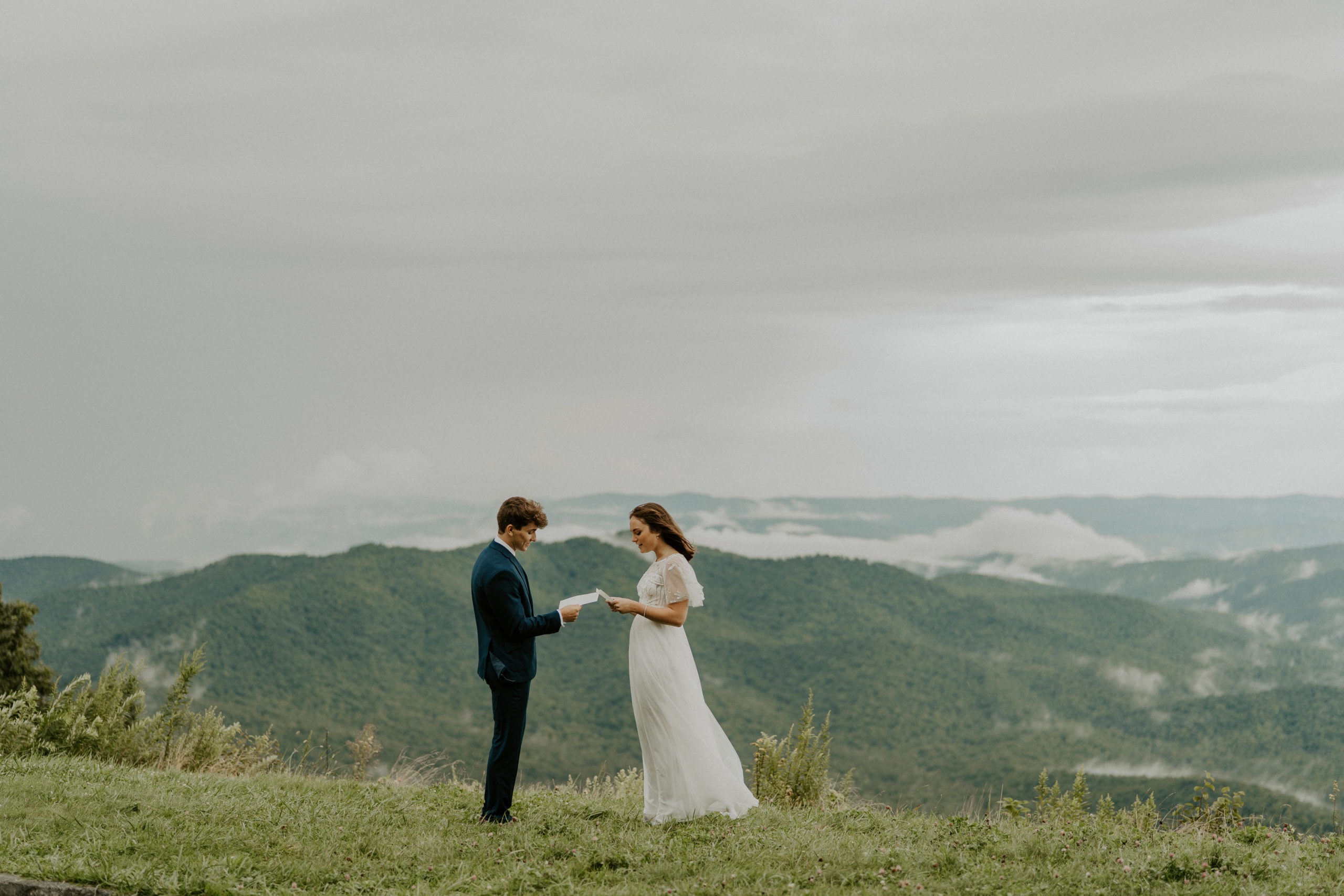 a man and woman exchanging vows with the Blue Ridge mountains in the background, they are facing each other and she is wearing a white wedding dress and he is in a blue suit.