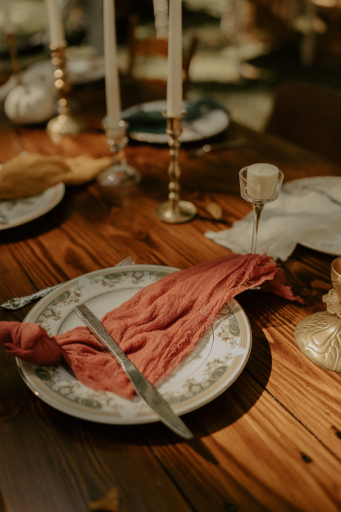 A dining table for the reception of a wedding. There are plates, candles, and napkins set up on a wooden table. The plates are ornate and vintage, as well as the rest of the items on the table. 