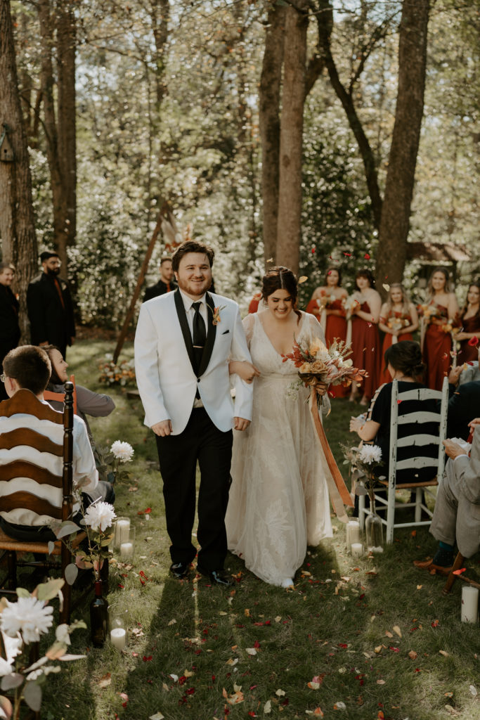 A couple in suit and dress, walking down the aisle right after getting married. There are trees in the background, and people in chairs in the foreground. The man is in a white suit, the woman is in an off-white dress and holding flowers in one hand. Her other hand is holding her husband's arm.