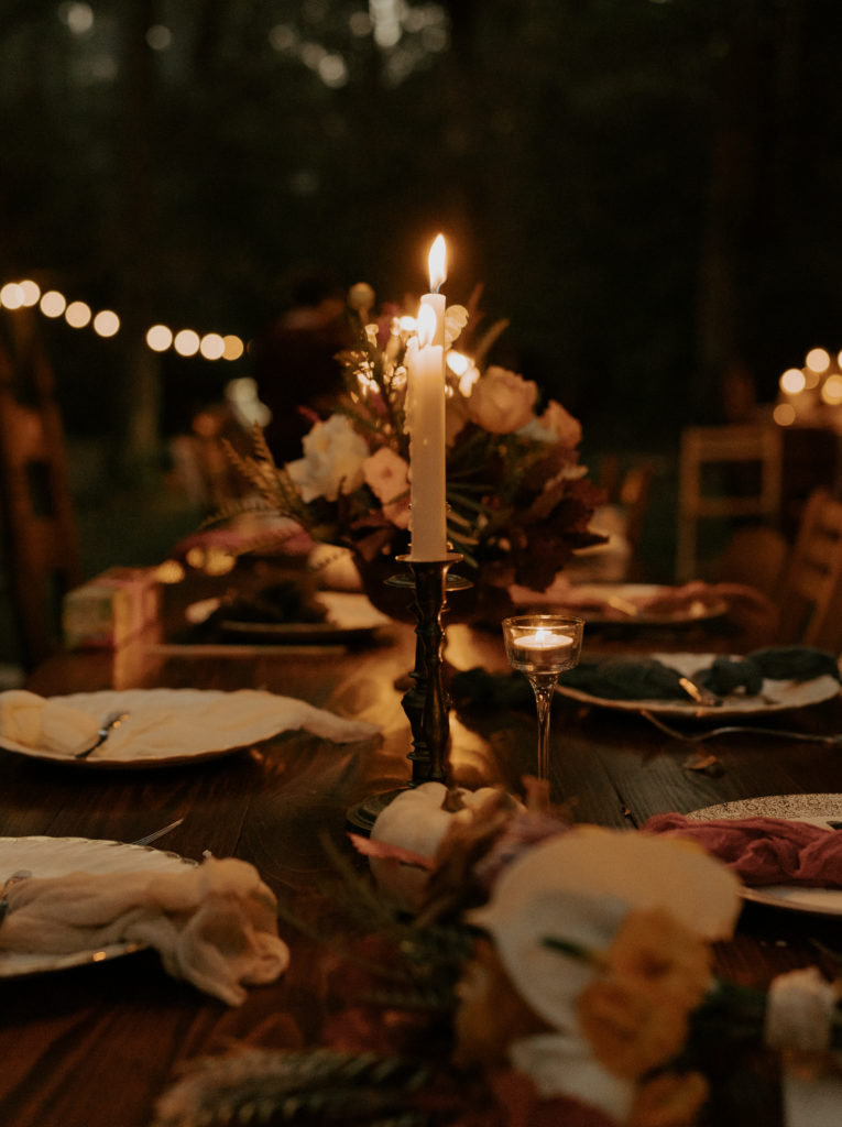 A dining table for the reception of a wedding. In the center there is a tall pillar candle burning, on the table below there are flowers, plates, and eating utensils. The image was taken in the late evening, so the warm candle light is illuminating the entire table. 