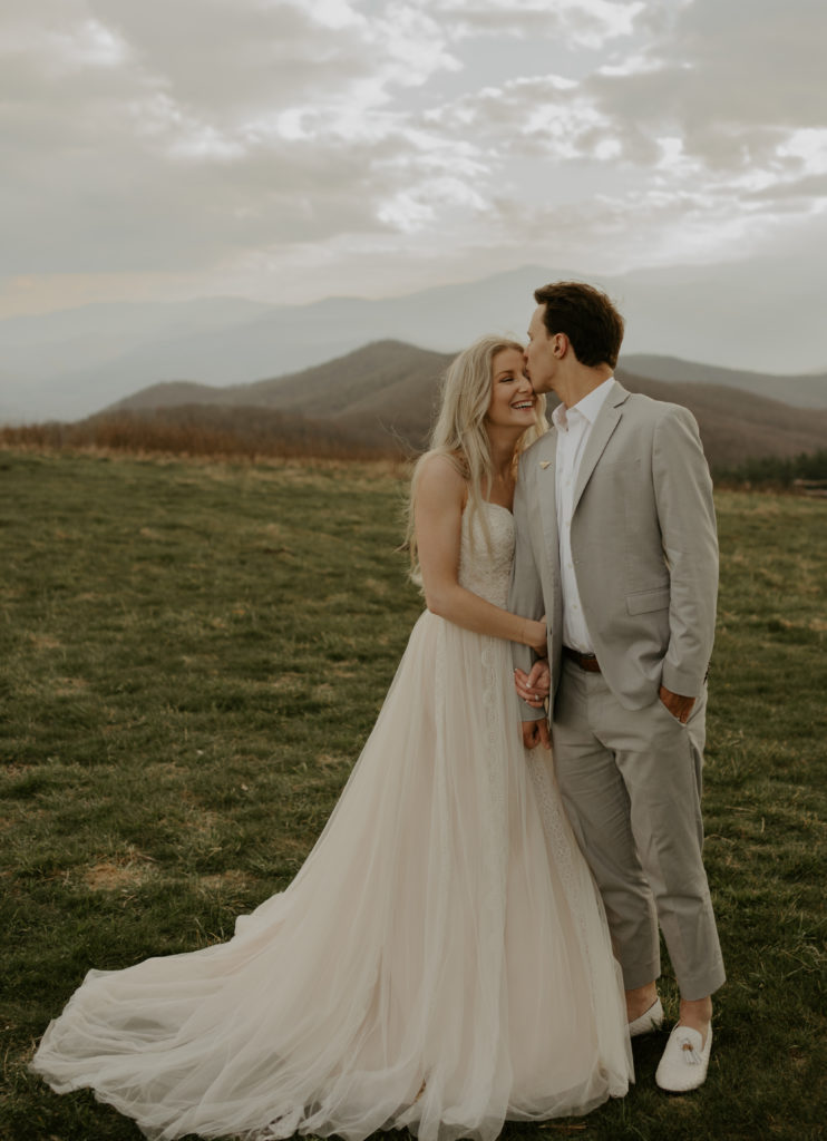 A couple eloping on top of Max Patch mountain in North Carolina. The man is kissing the woman on the forehead, and she is smiling. She is in a dress and he in a suit. In the background one can see mountains disappearing over the horizon.