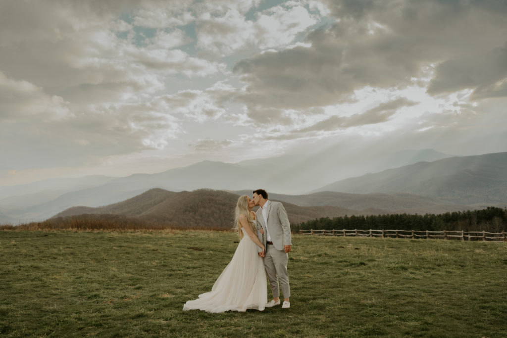A couple kissing on Max Patch mountain in North Carolina. One can see a large portion of the mountain, a fence in the background, and sweeping views of other mountains. She wears a long wedding dress, he a beige suit and pants. 