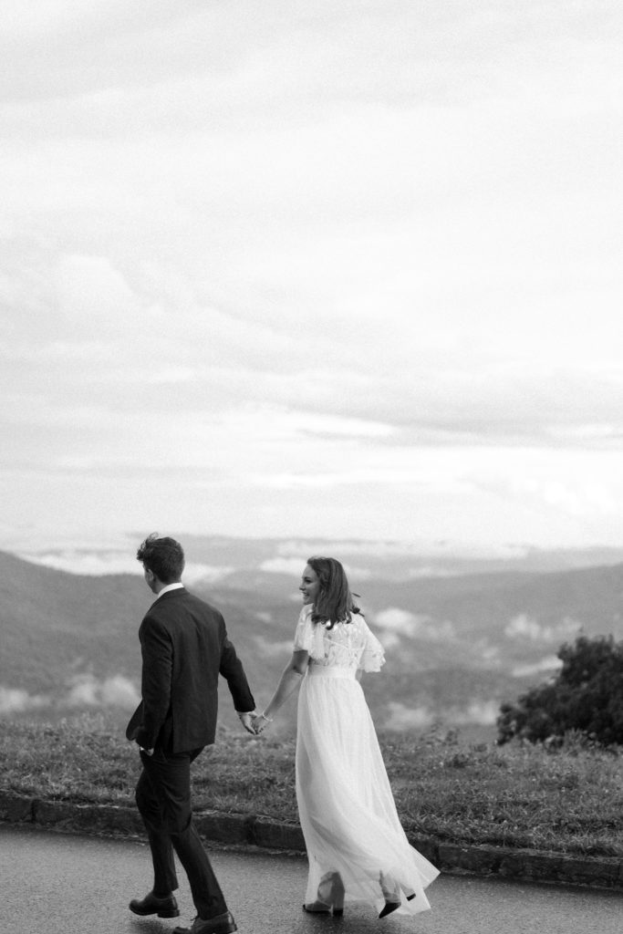 A black and white image of a couple walking on a mountain overlook at the Blue Ridge Parkway. She in a long wedding dress, he in a black suit. They walk away from the viewer facing the left side of the image. They are holding hands and sharing laughter.