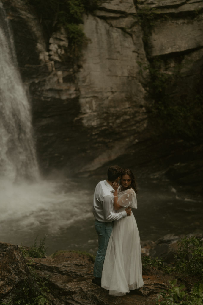 man and woman embracing in front of a waterfall. The woman is wearing a long, white wedding dress, and the man is wearing a white button up. There are rocks under their feet and a wall of rock in the background next to the waterfall.