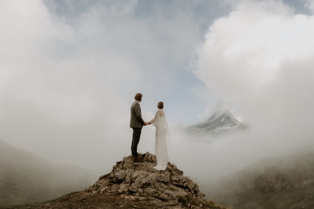 A woman and man eloping in Switzerland. They are standing on top of a mountain looking out at another mountain through the clouds.