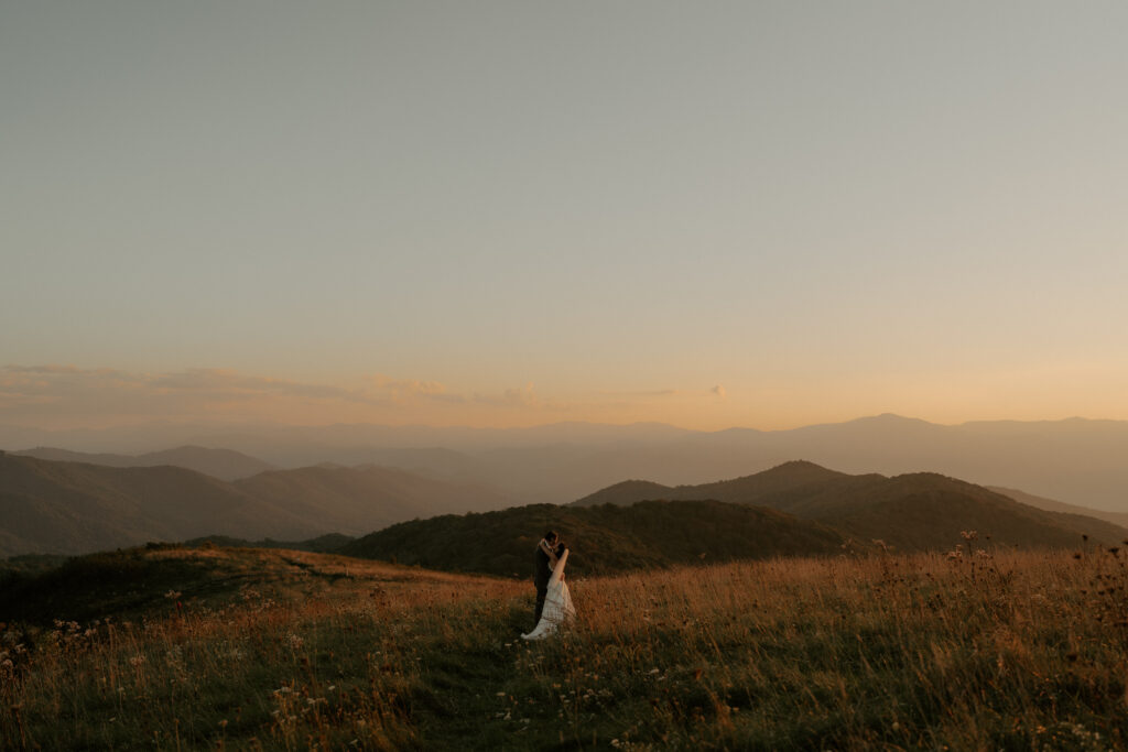 A man and a woman embracing and eloping in Asheville, North Carolina. The Blue Ridge mountains are behind them, and there are wildflowers all around them