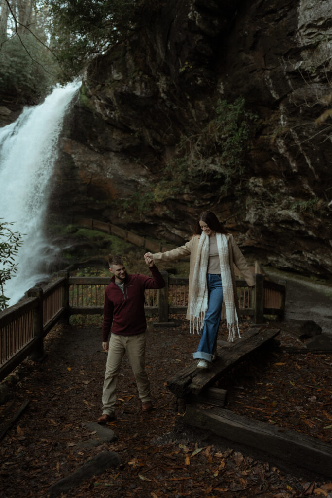 A couple stands together in stylish layered outfits, perfect for a cozy, seasonal look. The man wears a dark red sweater, cream pants, and brown shoes, while the woman pairs dark blue jeans with white sneakers, a tan shirt, a brown long coat, and a thick, long scarf. Their accessories, including earrings and a bracelet, add thoughtful details to the well-balanced outfit combination.