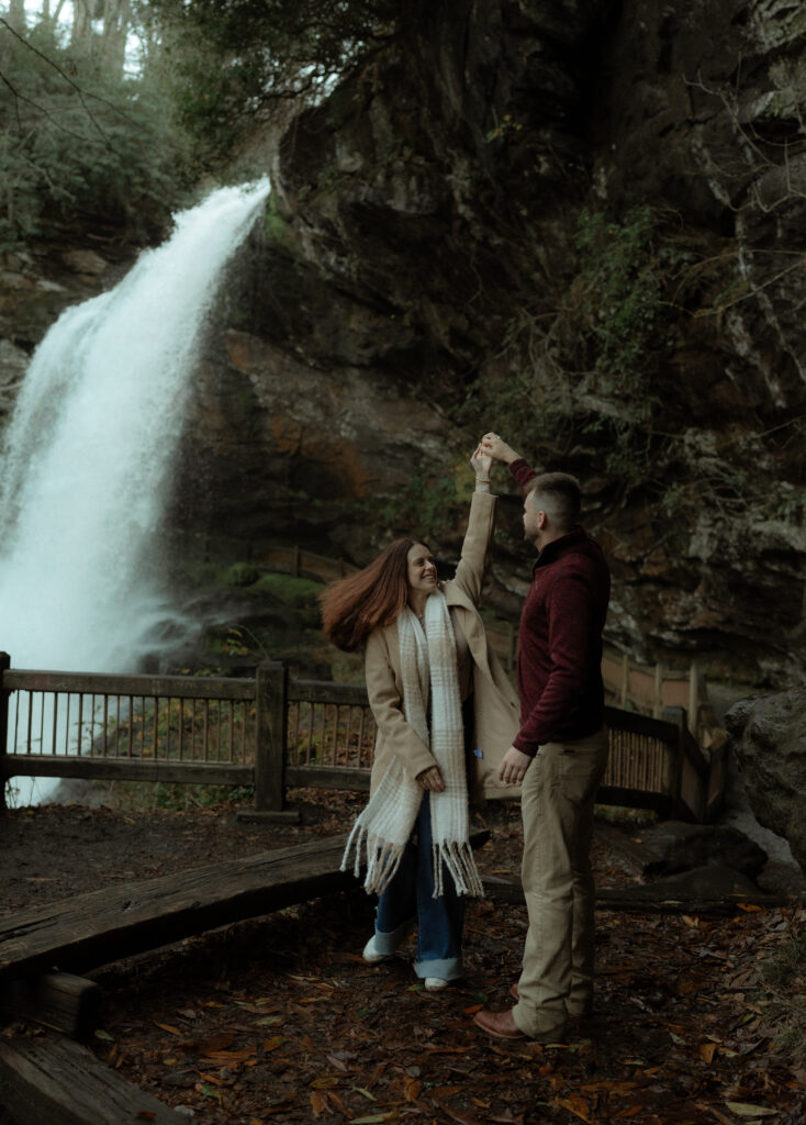 A couple dances in front of a waterfall as the man twirls the woman, causing her long scarf to flow behind her. Their layered outfits add texture and movement, complementing the natural beauty of the cascading water in the background.