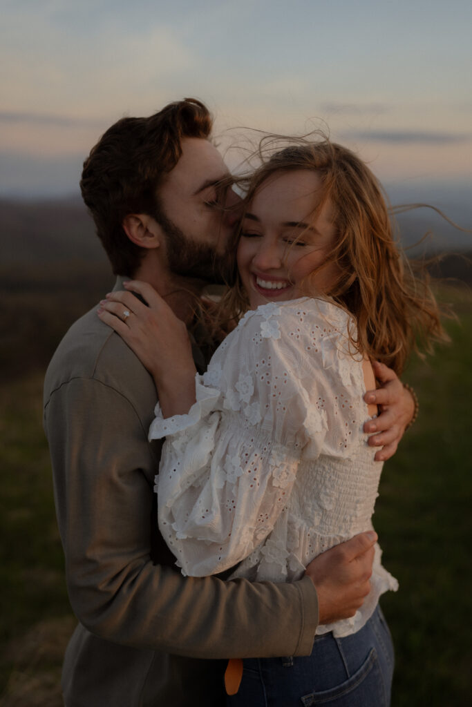 Close-up of a couple embracing as the man kisses the woman’s cheek. Her hair flows in the wind, adding movement to the moment. She wears a white flowy shirt with ruffles and floral details, blue jeans with a brown belt, and brown boots, while he is dressed in a muted dark green long-sleeve polo and khaki pants.