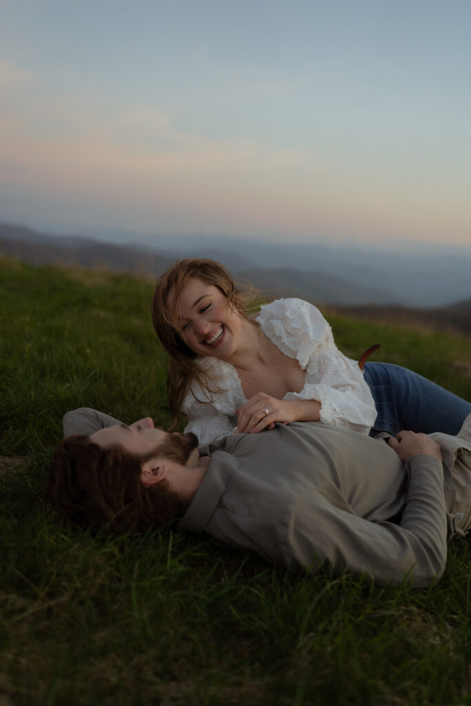 A couple lays together in a lush green field surrounded by mountains. The woman sits up slightly, gazing at the man with a soft expression. She wears a white flowy shirt with ruffles and floral details, blue jeans, and brown boots, while he is dressed in a muted dark green long-sleeve polo and khaki pants. The setting creates a peaceful and romantic atmosphere.