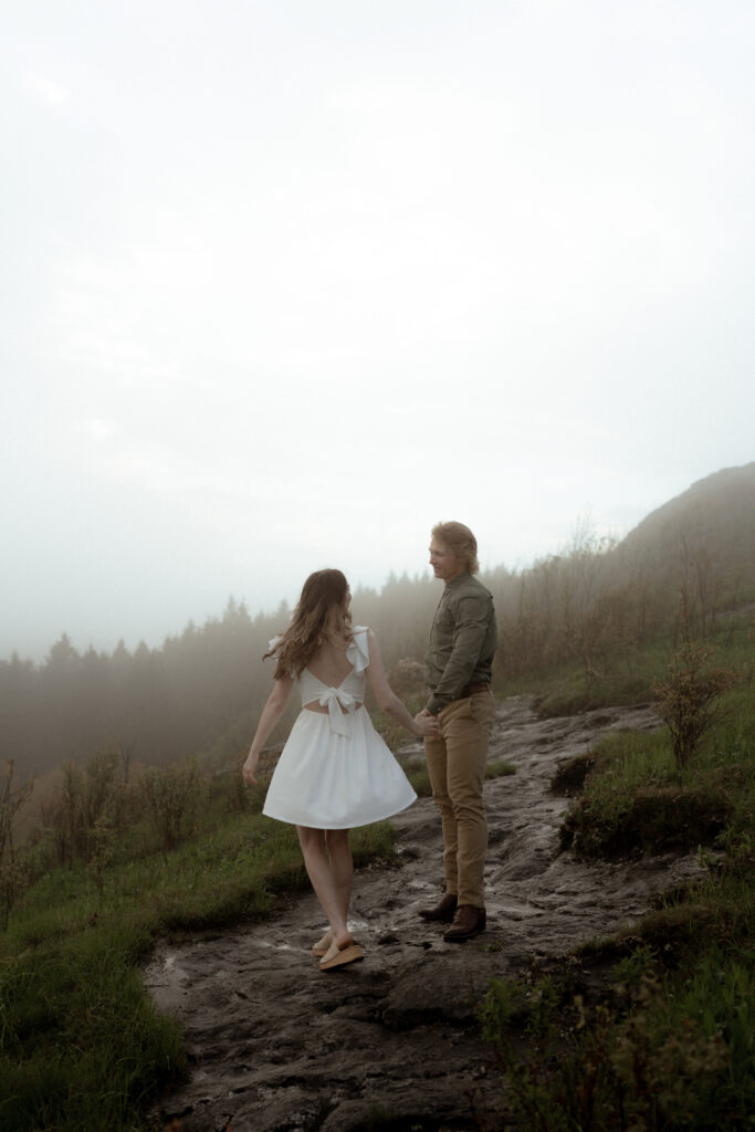 A couple dances outdoors as the man twirls the woman, her flowy white dress and hair moving gracefully with the motion. The natural setting, combined with their effortless movement, creates a romantic and carefree moment.