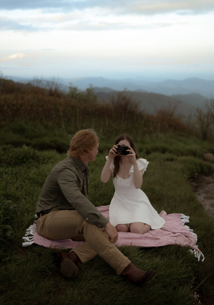 A couple sits on a picnic blanket surrounded by lush greenery and mountains. The woman, wearing a white short flowy dress with ruffled sleeves, holds a film camera and takes a photo of the man, who is dressed in a dark green button-up shirt, beige pants, and brown shoes. The scene captures a nostalgic and romantic outdoor moment.