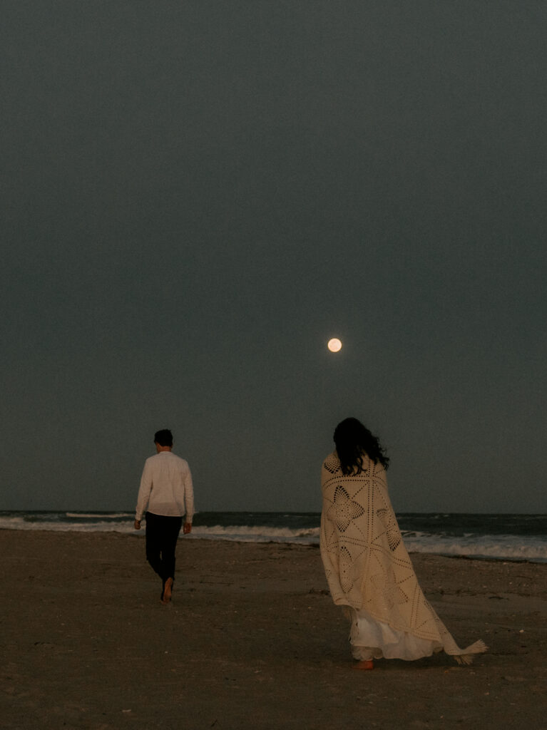 A couple walks along the beach, with the woman wrapped in an off-white blanket and the man leading slightly ahead in a white button-up shirt and black pants. The soft tones of their outfits complement the serene coastal setting, creating a peaceful and intimate moment.