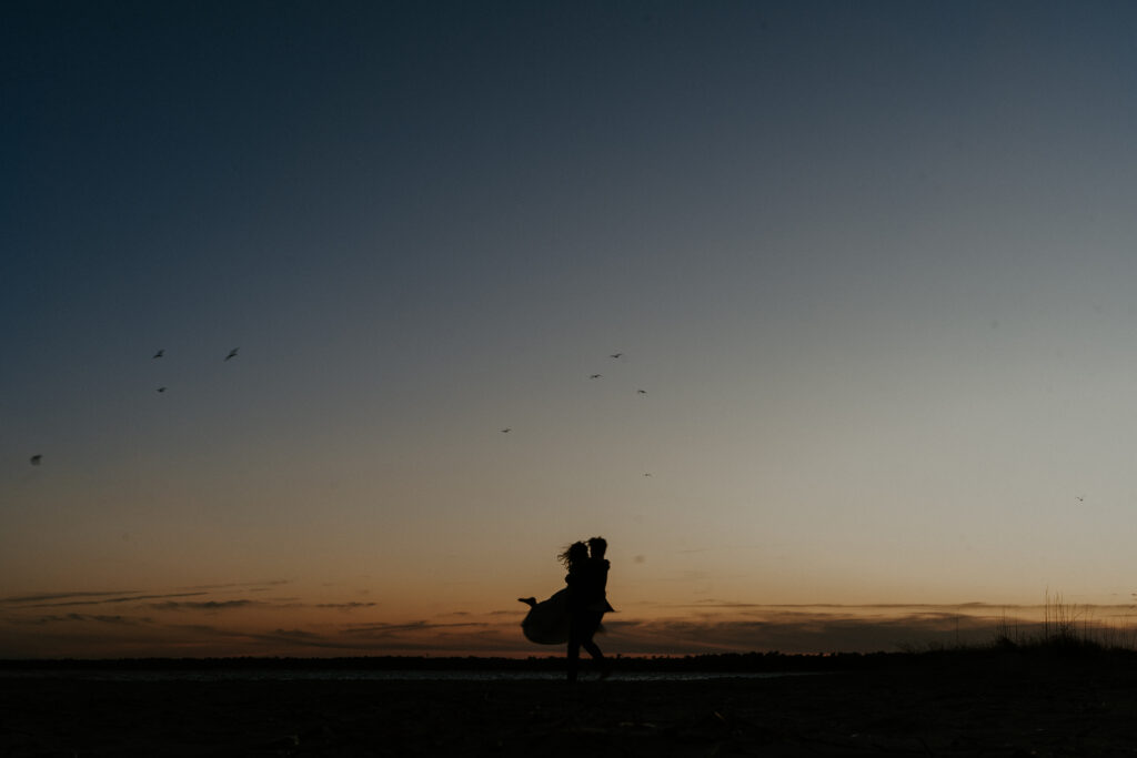 Silhouette of a couple as the man lifts and swings the woman around, her flowy dress billowing in motion. The dynamic movement and soft fabric create a striking, romantic scene against the backdrop of light and shadow.