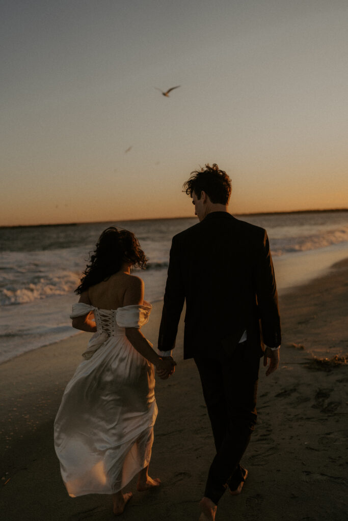 A couple walks hand in hand along the beach as birds fly in the distance. The woman’s silky dress catches the warm glow of the sunset, creating a soft, golden reflection. The man, dressed in a black suit, walks beside her, completing the romantic and serene scene