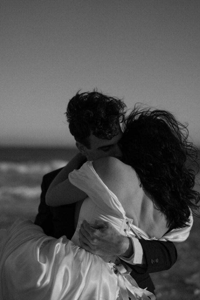 A couple shares a romantic moment on the beach as the man holds the woman in his arms. She wears a flowy white dress with off-the-shoulder sleeves, while he is dressed in a black suit with a white button-up shirt underneath. The soft ocean breeze adds movement to her dress, enhancing the dreamy, intimate feel of the scene.