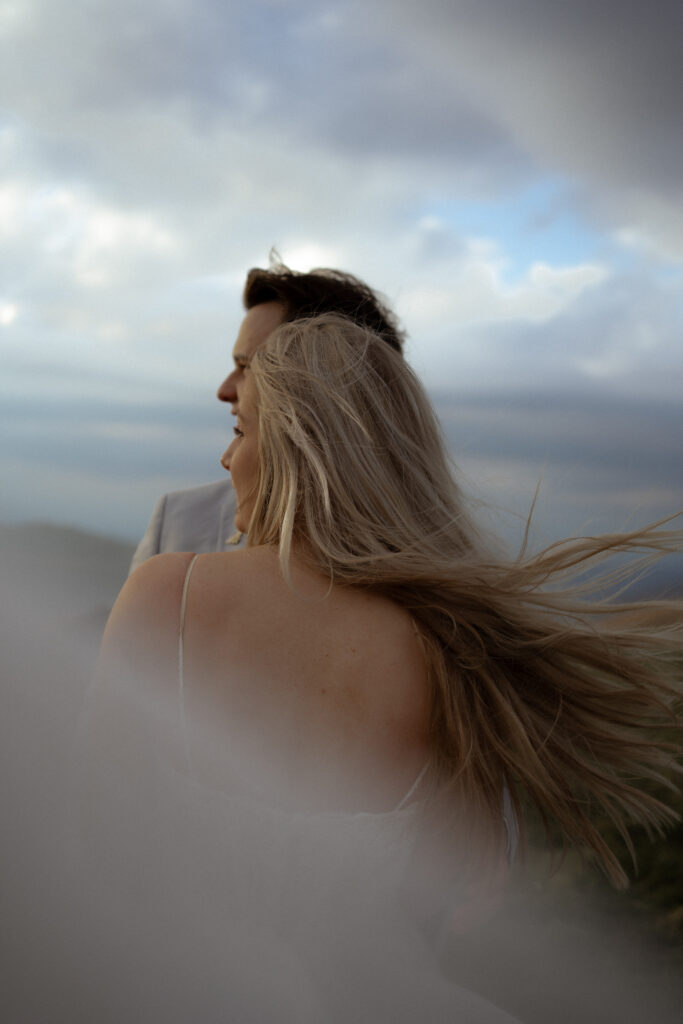 A couple embraces on Max Patch in North Carolina while gazing into the distance, the wind catching her hair and dress in a cinematic, dreamlike moment