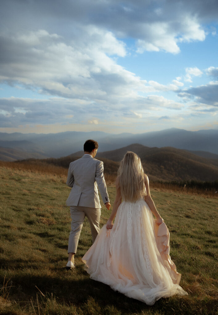 A couple walks hand in hand along a mountain ridge, surrounded by dramatic clouds and sunbeams stretching over miles of rolling peaks.