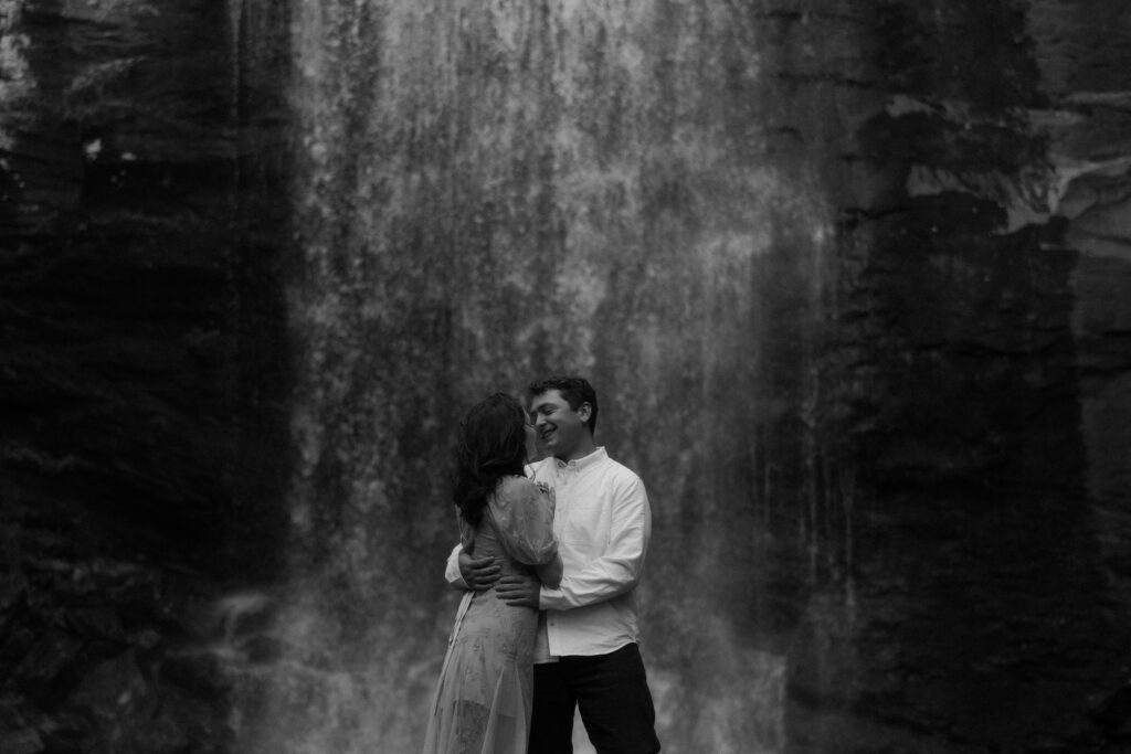 Black and white portrait of an eloping couple standing together in front of a waterfall near Asheville, North Carolina. A timeless moment showcasing the natural beauty that can be part of an elopement experience and budget.