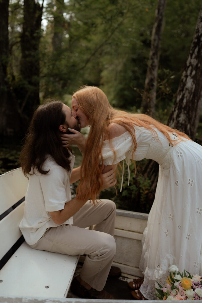 A couple shares a kiss on a boat as she leans in toward him, surrounded by peaceful water and lush trees in a romantic outdoor setting.