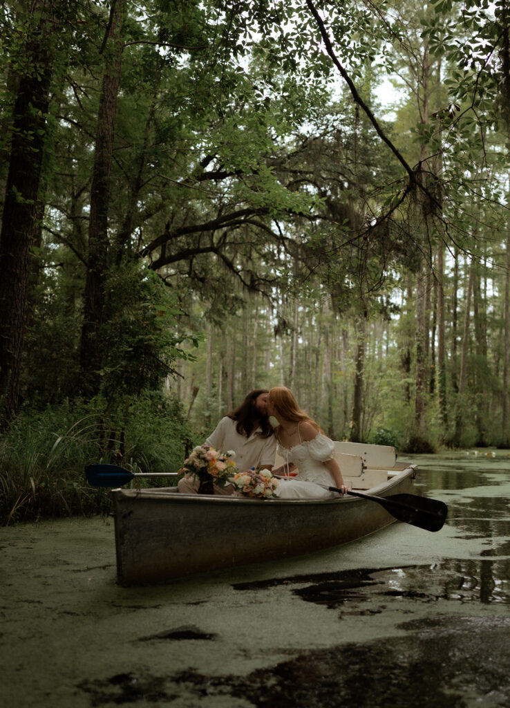 A couple shares a kiss on a boat beneath a canopy of trees, creating a romantic and serene wedding day moment on the water.