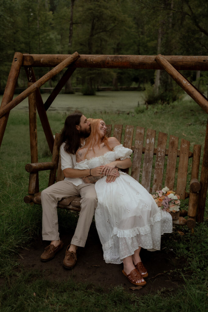 A couple shares a quiet moment on a wooden swing, as he kisses her cheek and she leans into him, surrounded by lush greenery and a bouquet nearby.