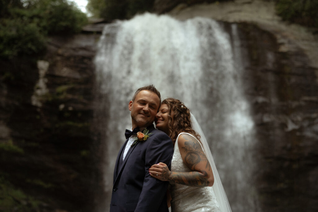 A couple in wedding attire stands in front of Looking Glass Falls in North Carolina, as she hugs him from behind and he smiles back at her.