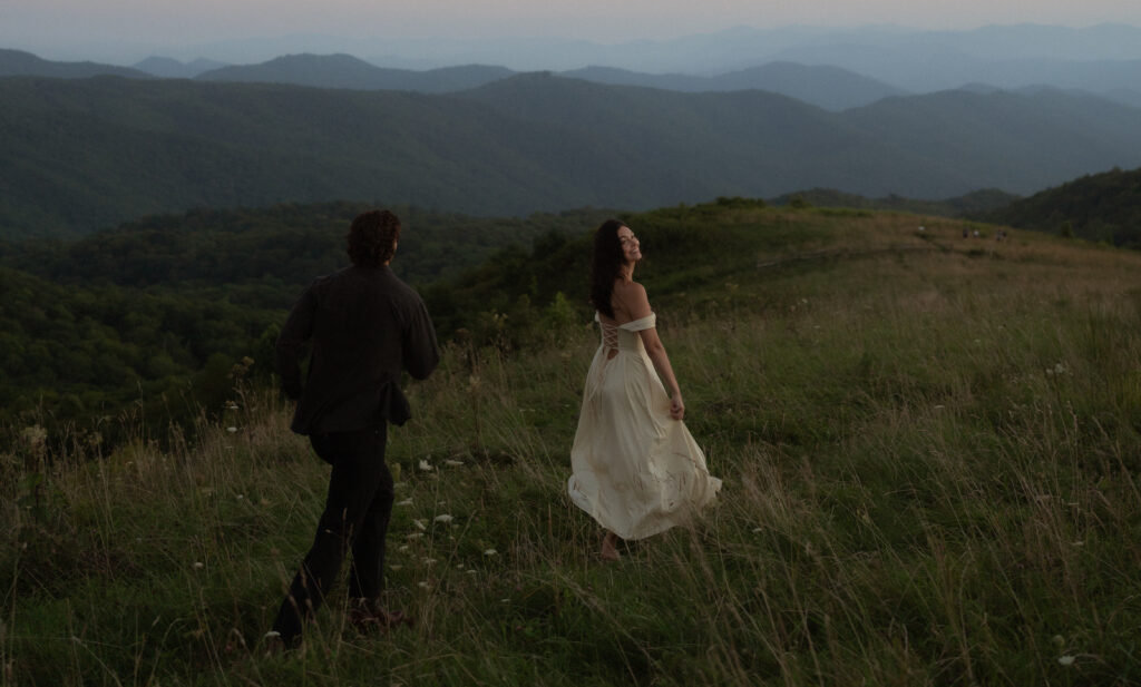 A couple runs across a field during blue hour, their silhouettes standing out against the soft evening light. The woman wears a pale yellow flowy dress with off-the-shoulder sleeves, while the man is dressed in black jeans and a black jacket. The contrast between their outfits and the moody sky adds depth and romance to the scene.