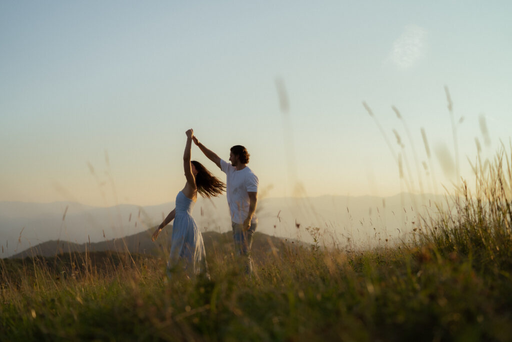 A couple dances in an open field as the woman's flowy light blue dress and hair move with the motion. She wears a sleeveless, airy dress, while the man, dressed in a white t-shirt and dark blue jeans, guides her in the dance. The golden sunlight and surrounding greenery enhance the dreamy, romantic feel of the moment.