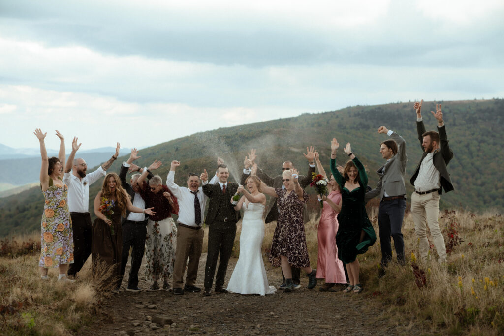All of the guests and the bride and groom popping a bottle of champagne with the hills in the background
