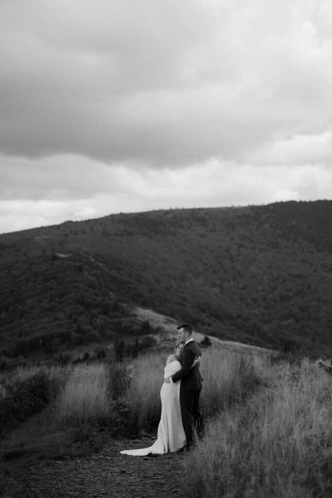 A black and white photo of a married couple embracing surrounded by mountains and wildflowers